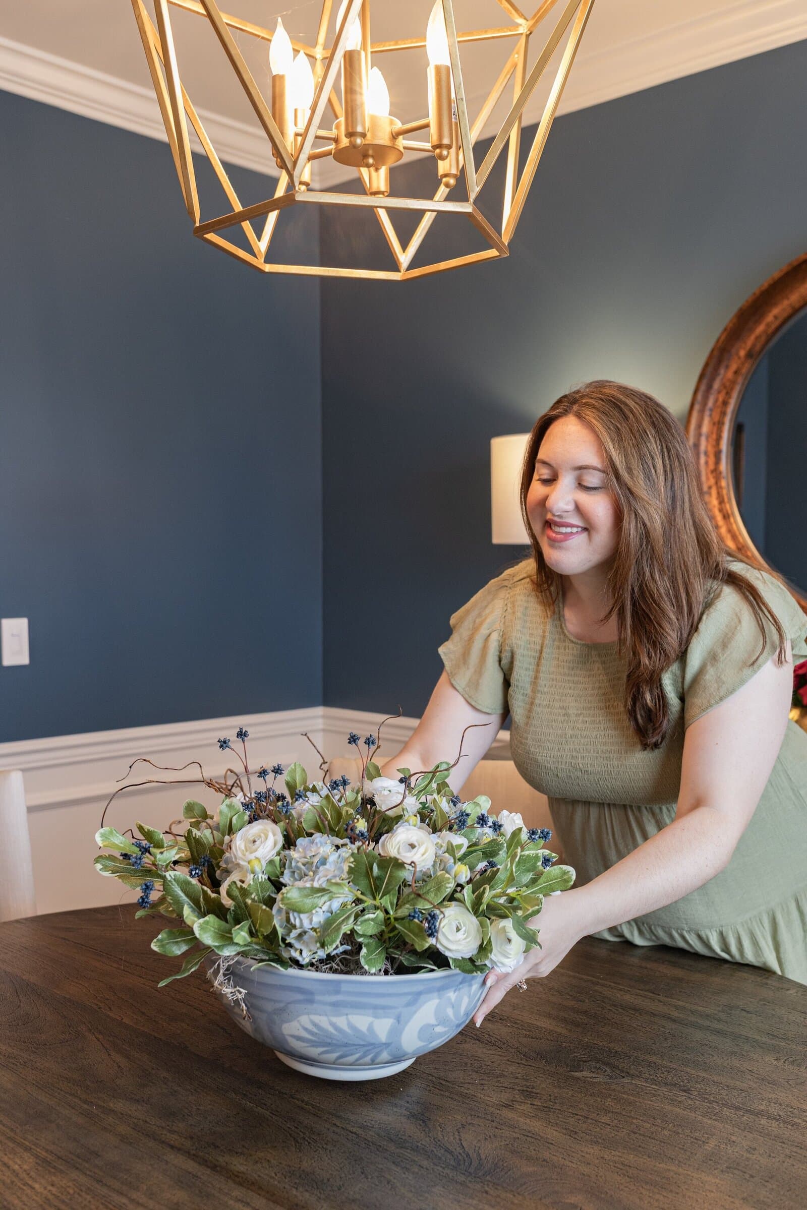 A navy dining room with white wainscoting, a gold geometric lantern over the table, and a low arrangement of white roses and hydrangea in a blue and white bowl