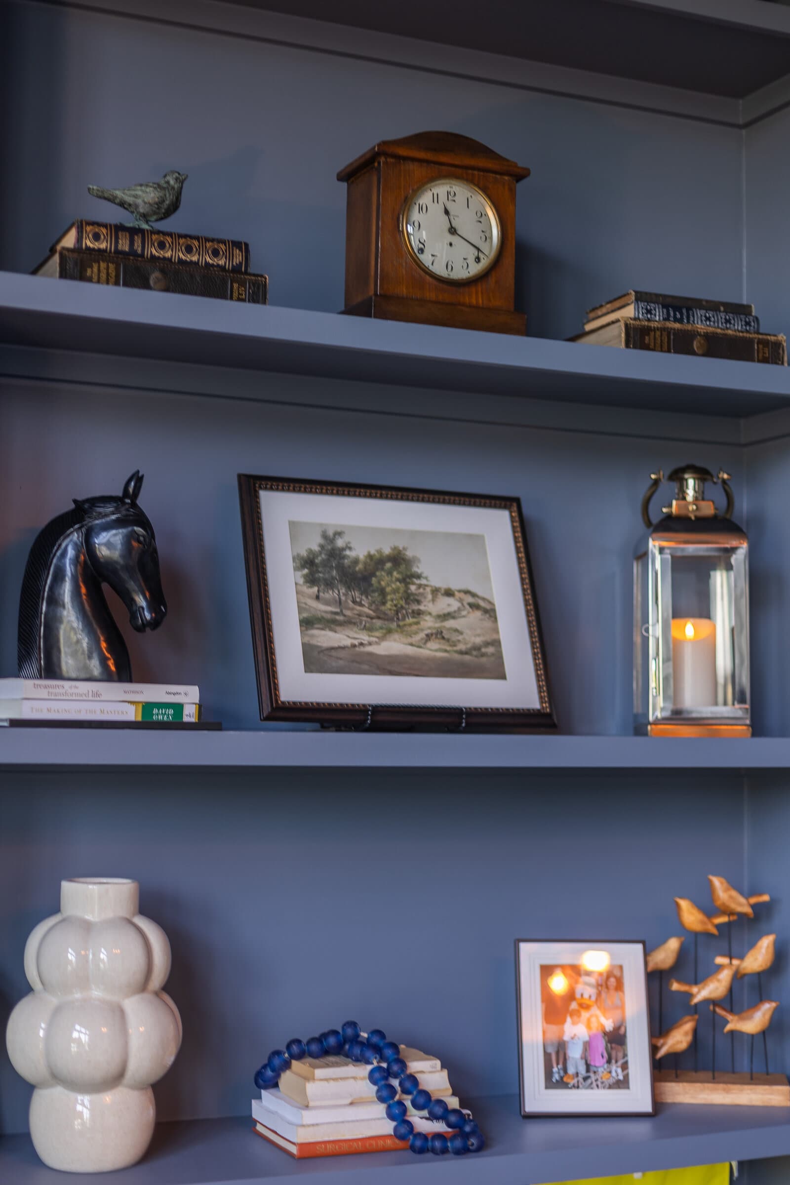 A close view of blue-grey shelves holding a bronze horse head, a framed landscape watercolor, a brass lantern with a lit candle and stacked antique books