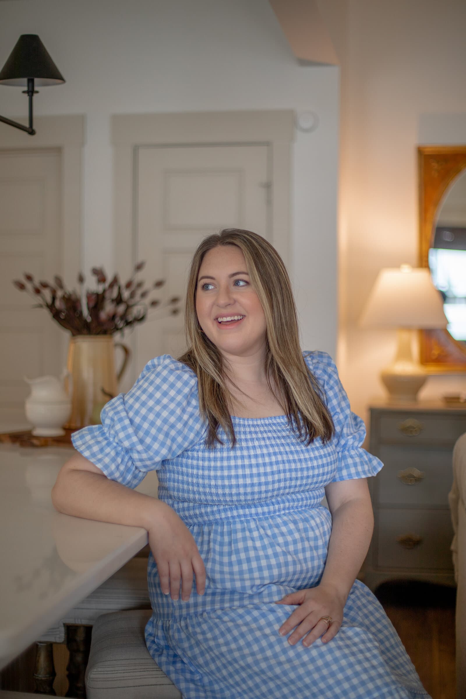 Kayla laughing, seated at a marble counter, with dried florals in a brass pitcher and a lamp on an antique chest behind her