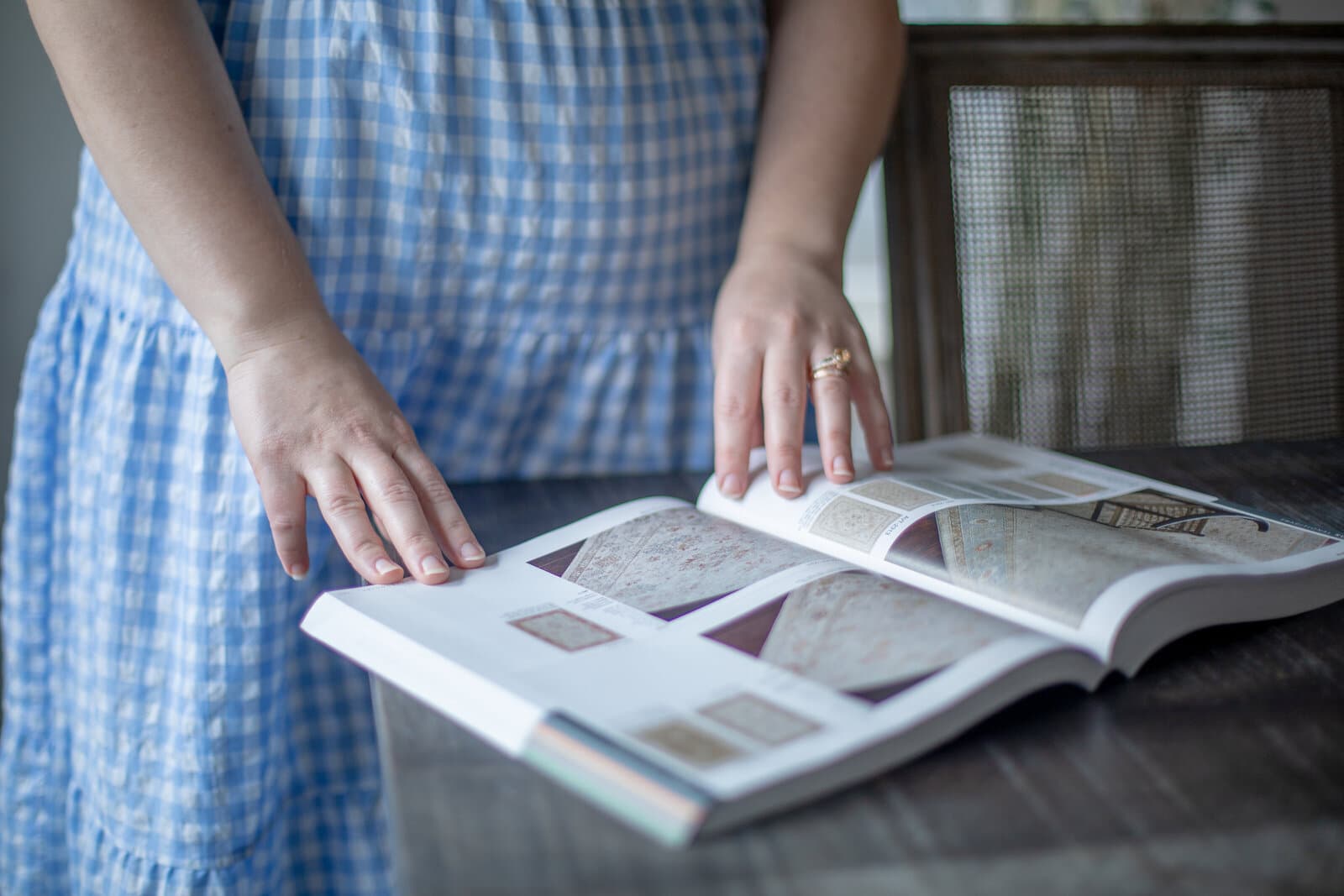 Hands resting on an open rug catalog spread across a dark wood table, beside a caned chair back