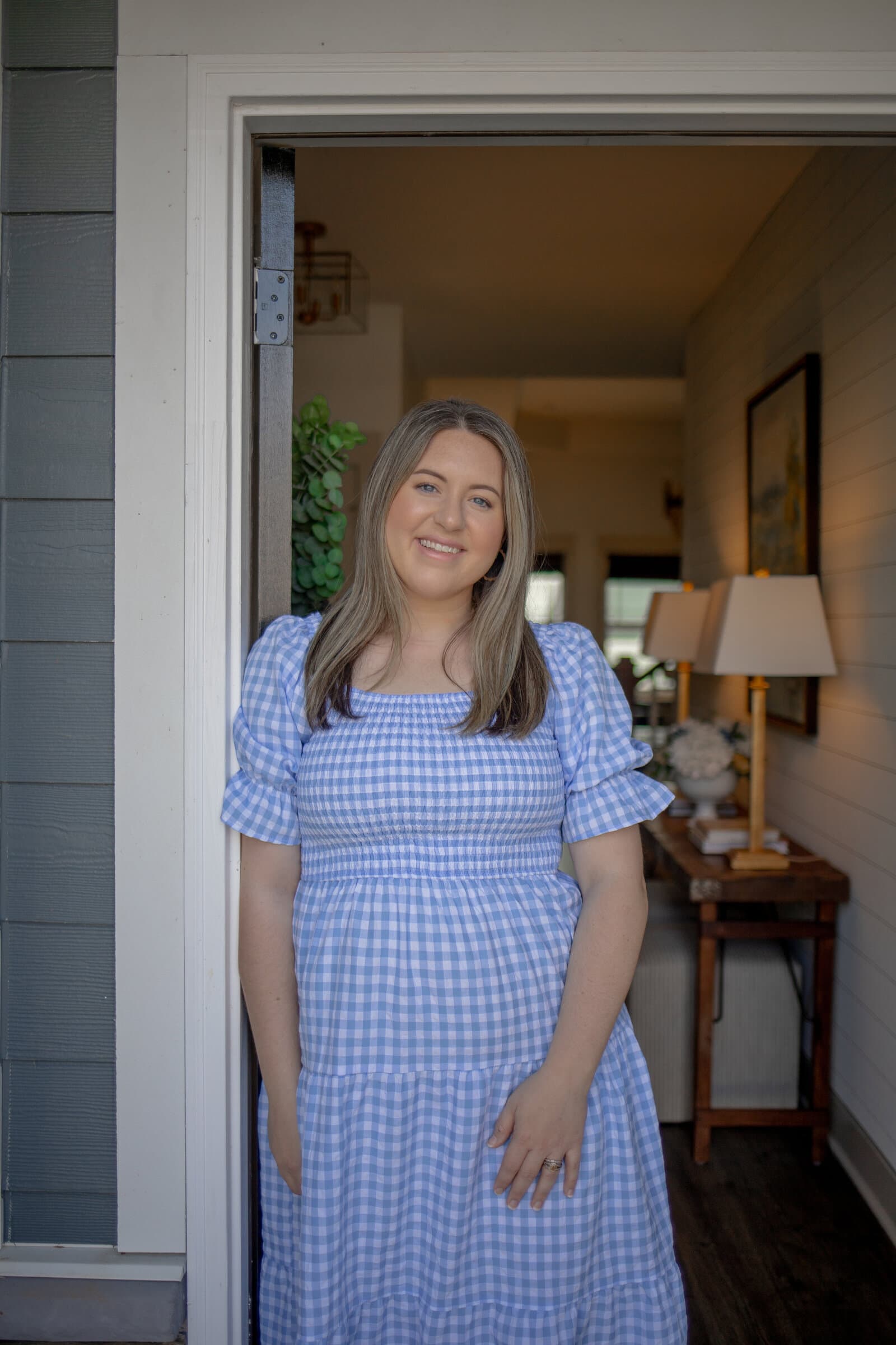 Kayla standing in the open doorway of a home, in a blue gingham dress, with a lamp-lit entry table visible behind her
