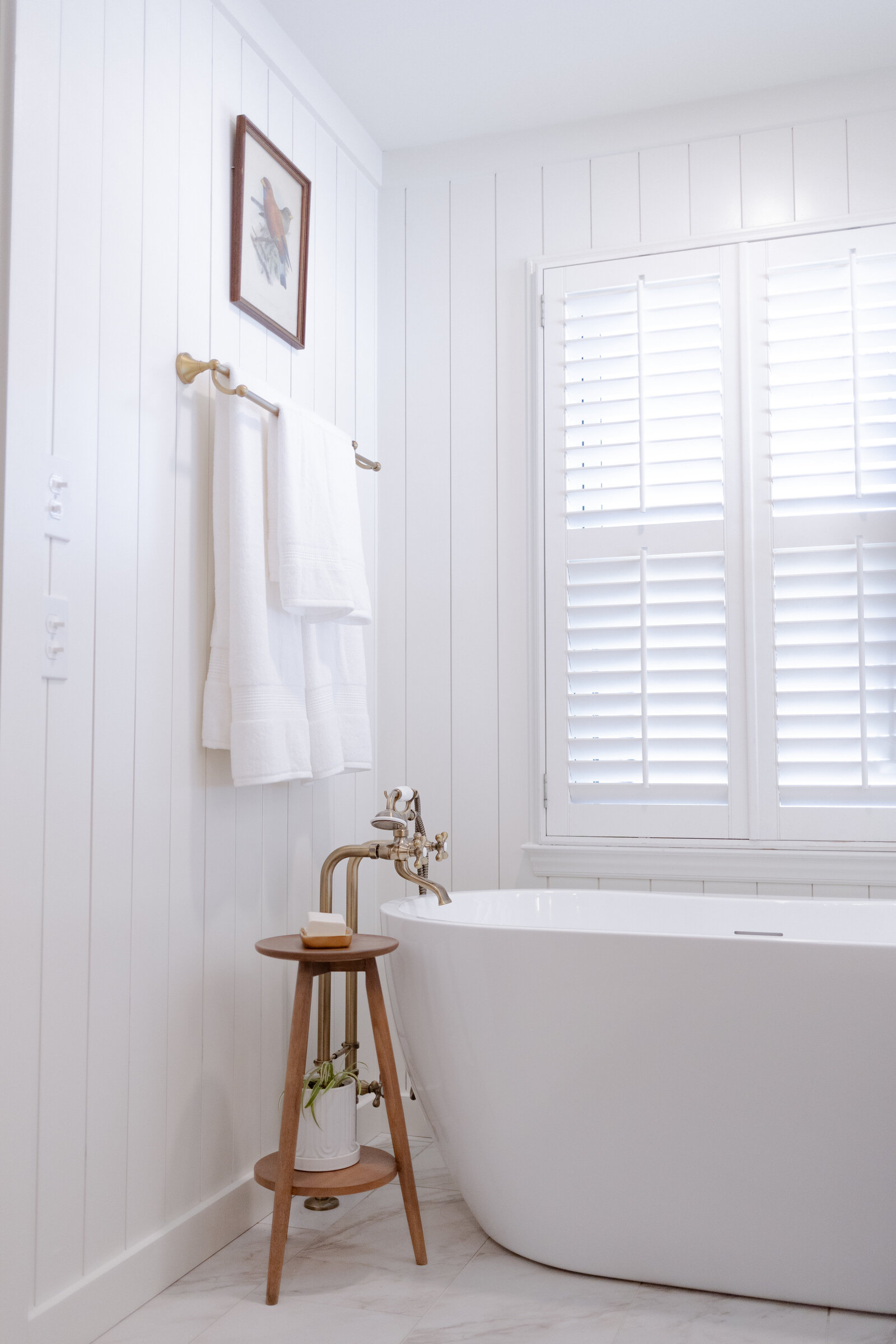A white bathroom with plantation shutters, a freestanding tub and a two-tier wooden table holding a soap dish
