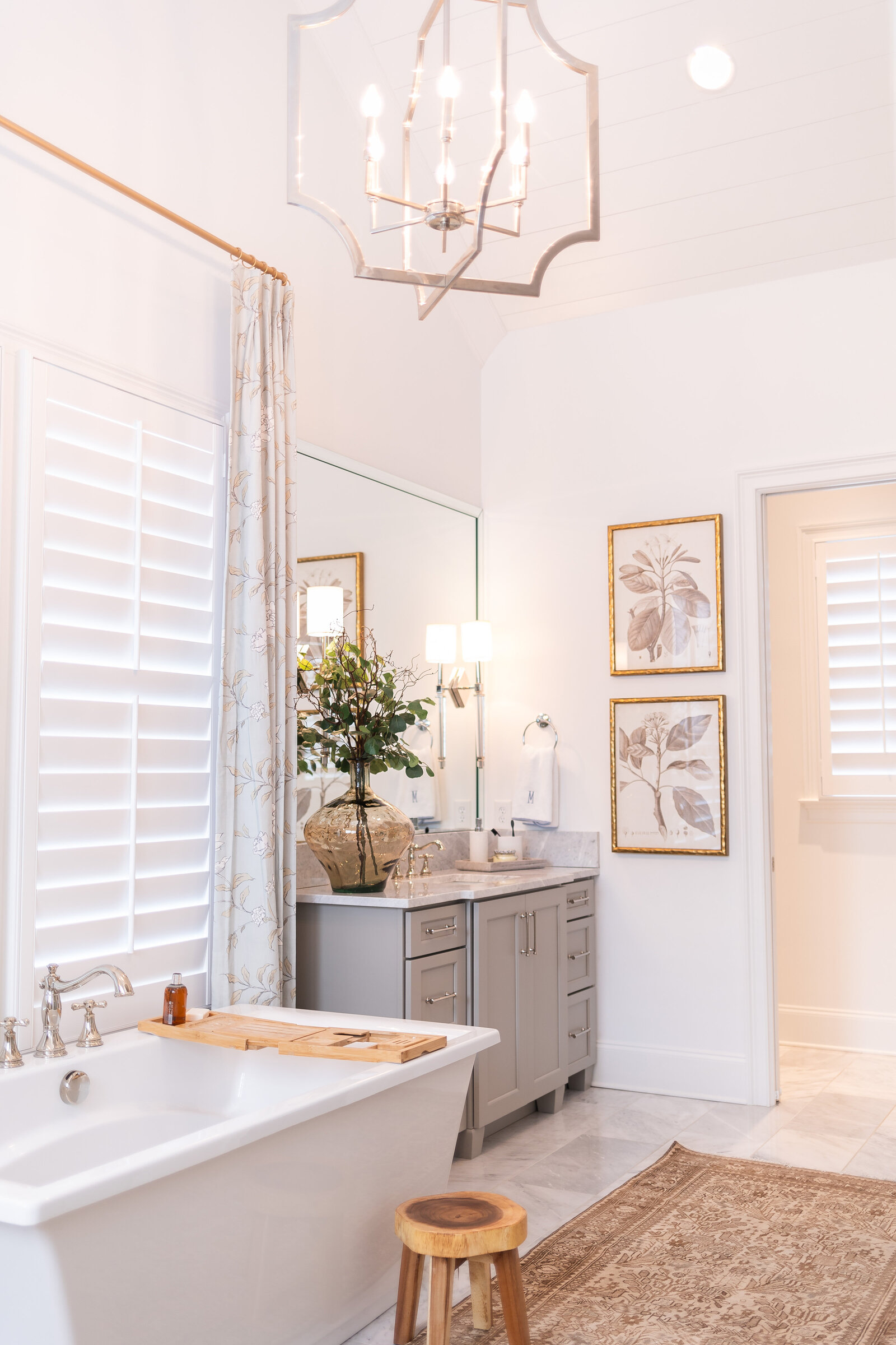 A primary bathroom under a vaulted shiplap ceiling, with a silver lantern chandelier, a freestanding tub and gold-framed botanical prints