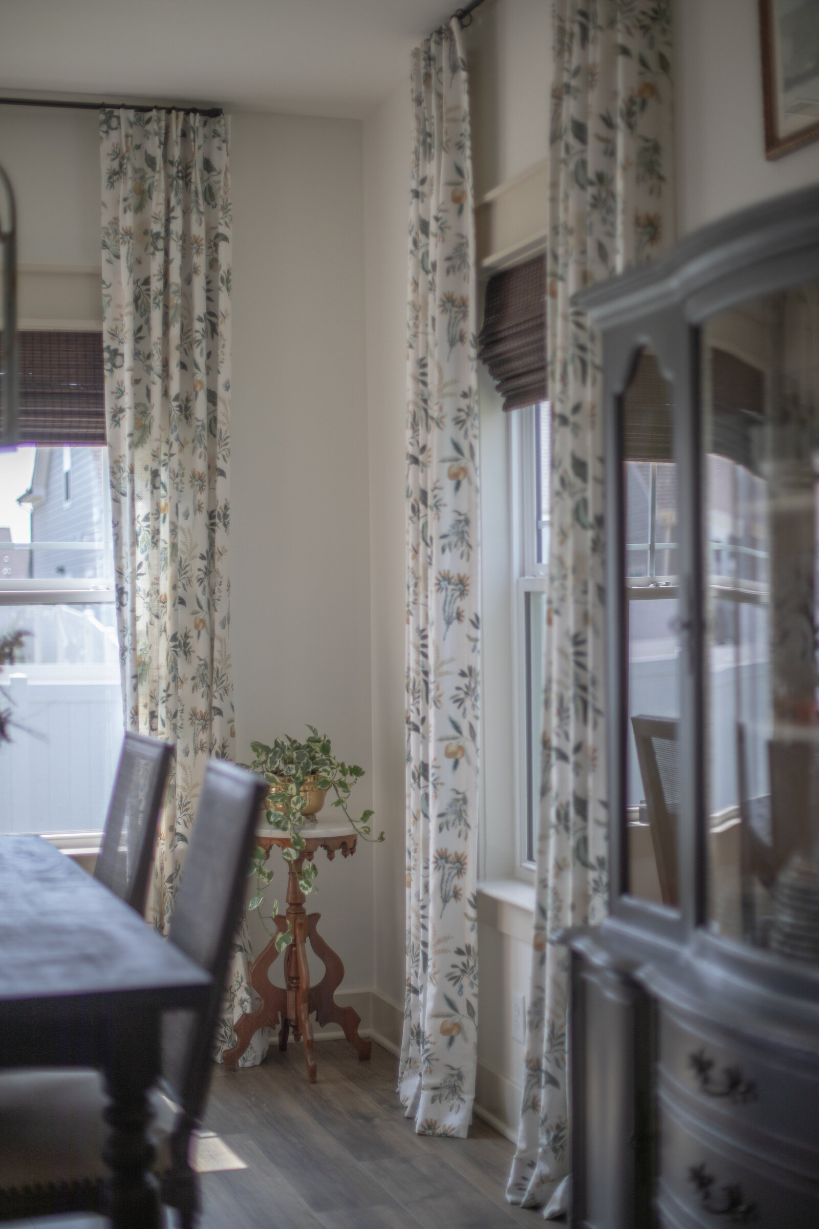A dining room with floral patterned drapery over woven shades, an antique marble-top table and a painted china cabinet