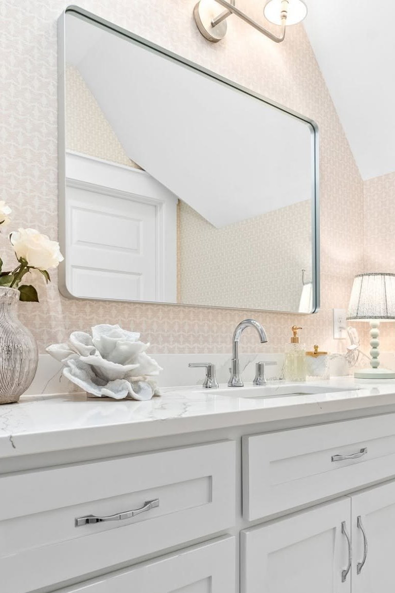 A bathroom vanity in pink patterned wallpaper, with a rounded rectangular mirror, a chrome sconce and a marble counter