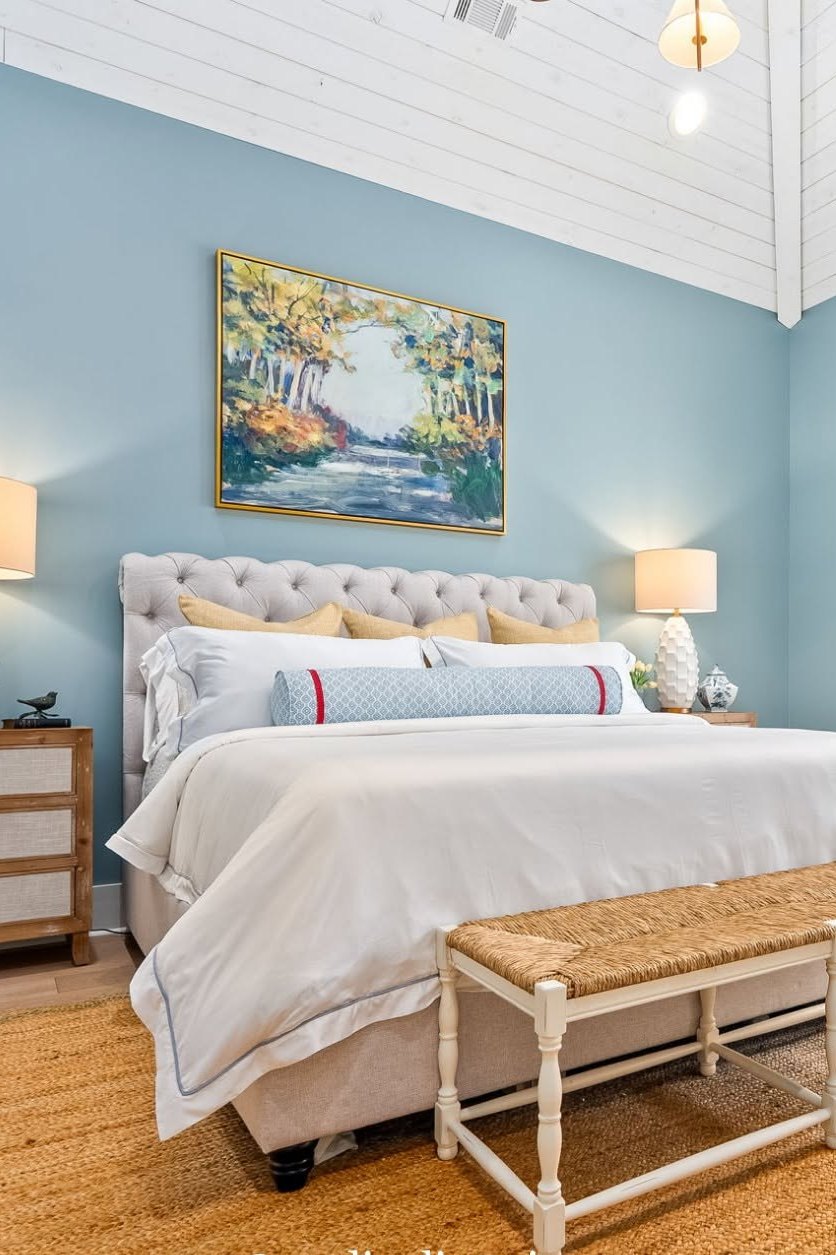 A blue bedroom with a tufted upholstered headboard, layered white bedding and a woven bench at the foot of the bed