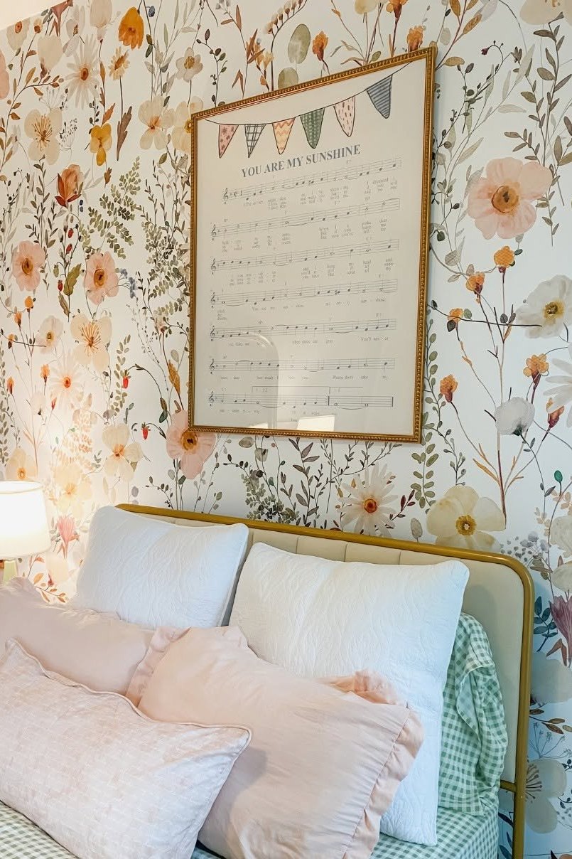 A bedroom in wildflower wallpaper with a brass bedstead, pink and green gingham bedding and a framed sheet music print