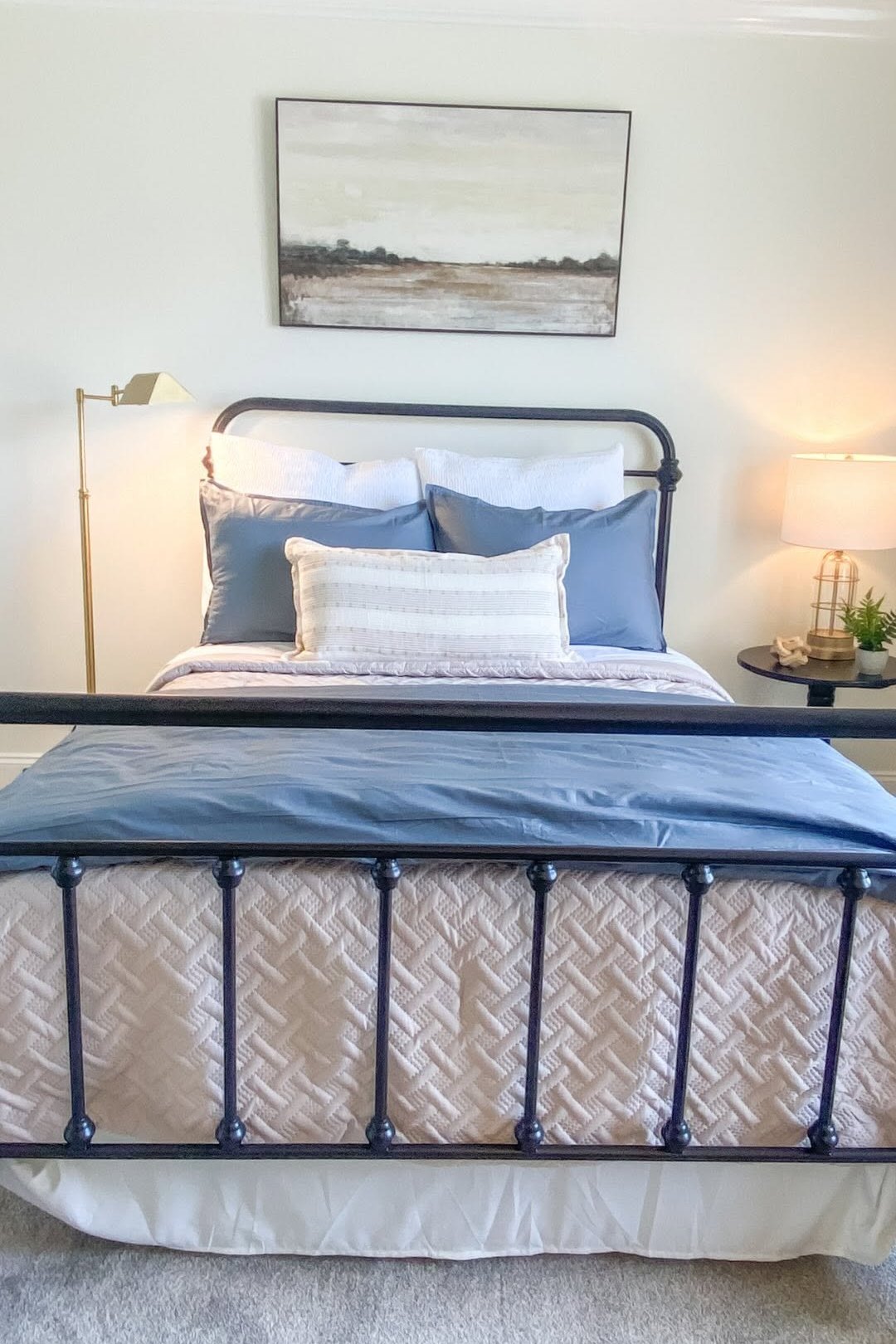 A bedroom with a black iron bedstead, blue and cream layered bedding and a brass swing-arm floor lamp
