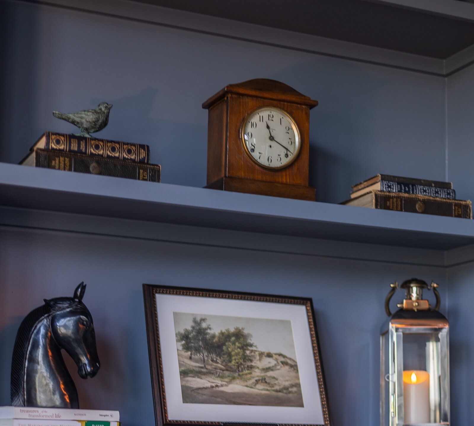Blue-grey built-in shelves holding an antique mantel clock, a verdigris bird on leather-bound books, a bronze horse head and a brass lantern with a lit candle